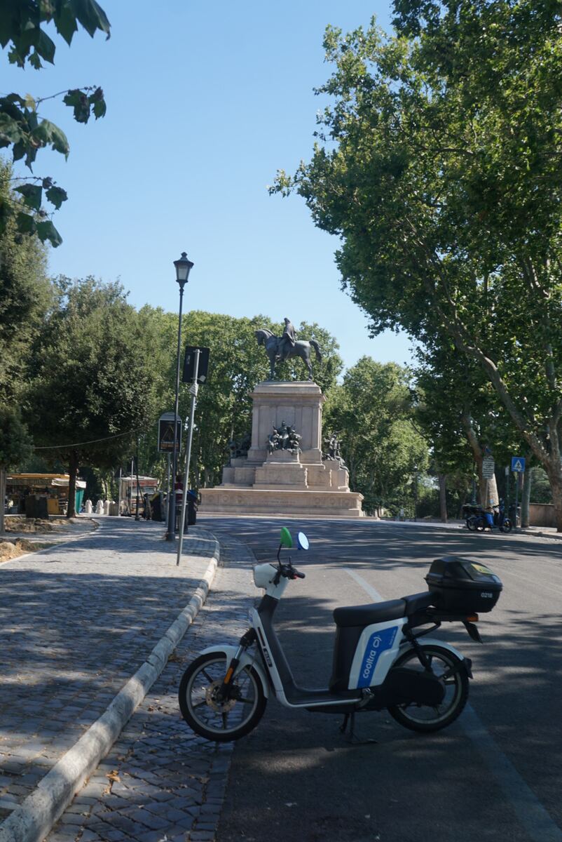 Een witte en blauwe scooter staat geparkeerd aan de rechterkant van een weg, met een geplaveide stoep aan de linkerkant. Op de achtergrond is een groot bronzen ruiterstandbeeld op een stenen sokkel te zien, omringd door veel groen en bomen onder een helderblauwe lucht. Er is ook een lantaarnpaal en enkele verkeersborden zichtbaar.