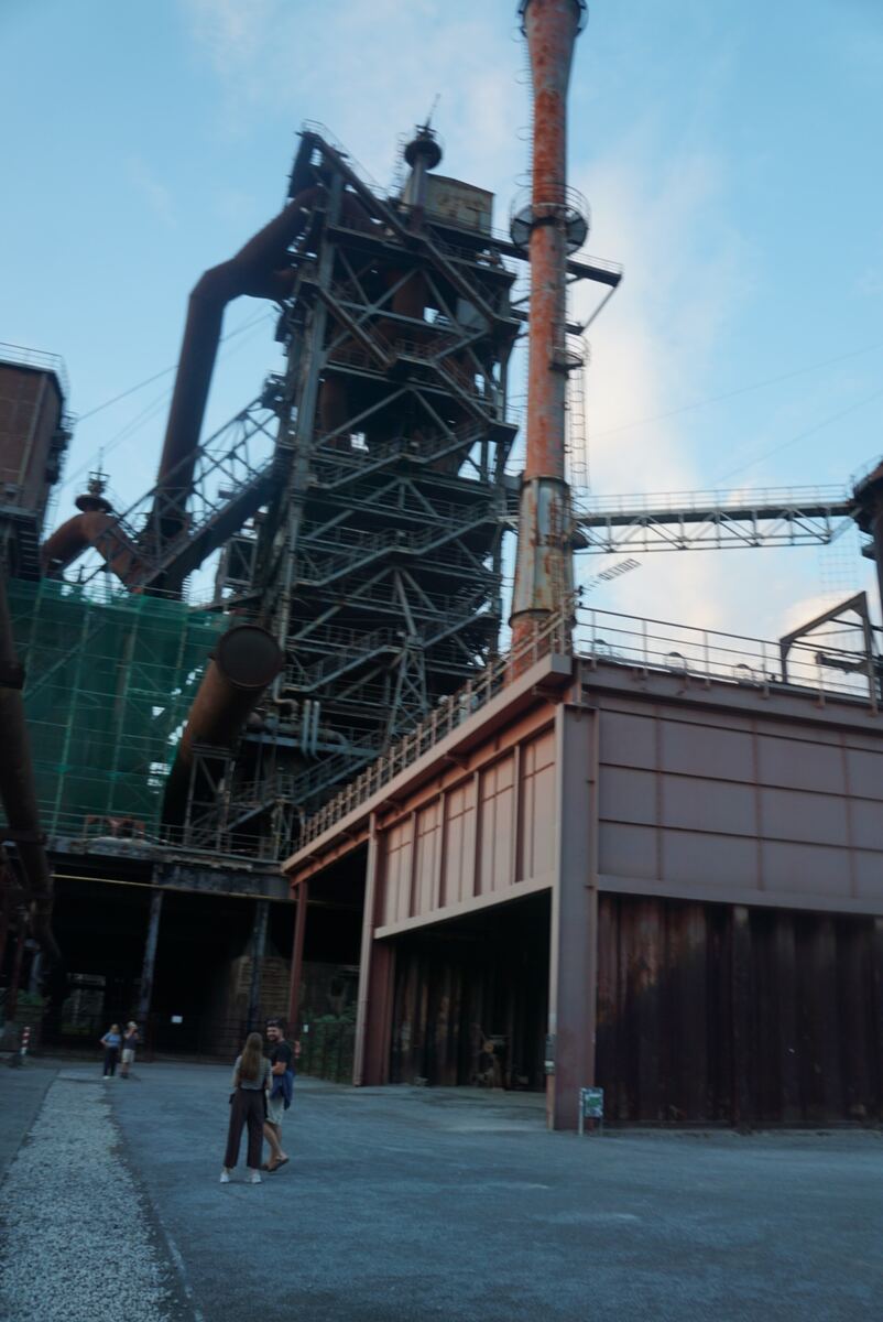 We zien een uitgestrekt voormalig industrieel complex, waarschijnlijk een hoogoveninstallatie, onder een deels bewolkte blauwe lucht. Grote roestige staalconstructies, pijpen en een hoge, slanke schoorsteen domineren het beeld. Links staat een massieve constructie met meerdere platforms en trappen. Op de voorgrond wandelen enkele mensen op een grindpad tussen de gebouwen door. De structuren tonen tekenen van verval en roest.