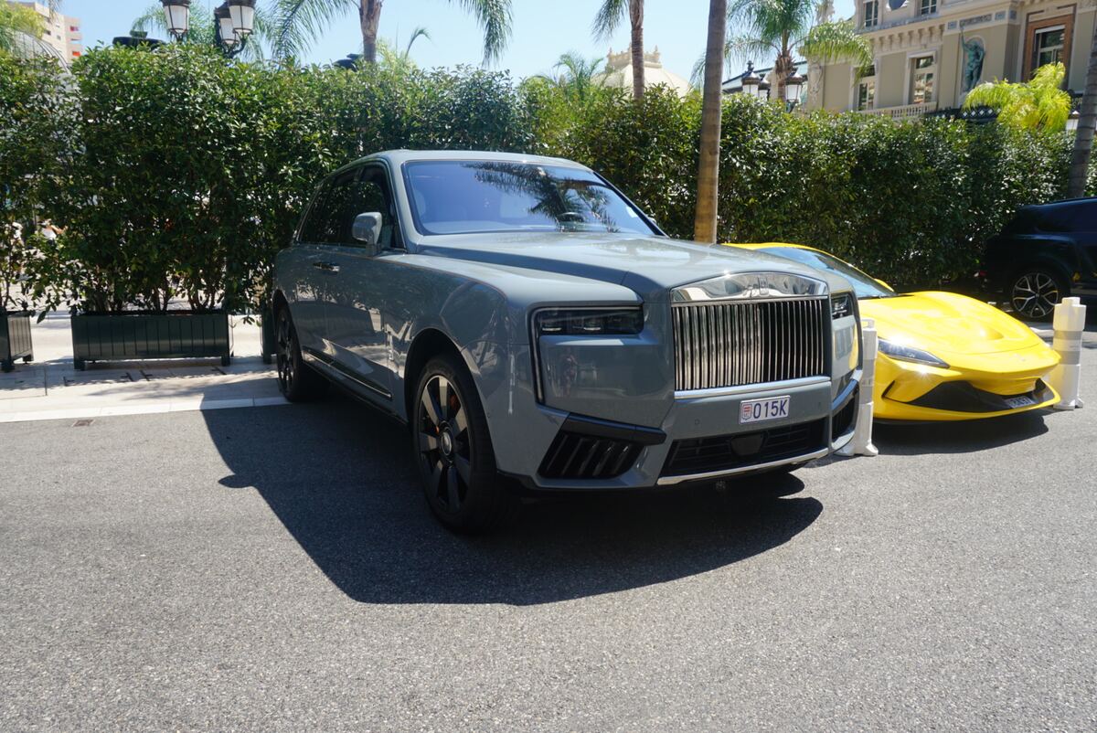 Een grijze Rolls-Royce Cullinan SUV staat geparkeerd op een geasfalteerde weg, met een felgele sportwagen deels zichtbaar erachter. Op de achtergrond zijn groene heggen, palmbomen en een klassiek ogend gebouw te zien onder een heldere, blauwe lucht.