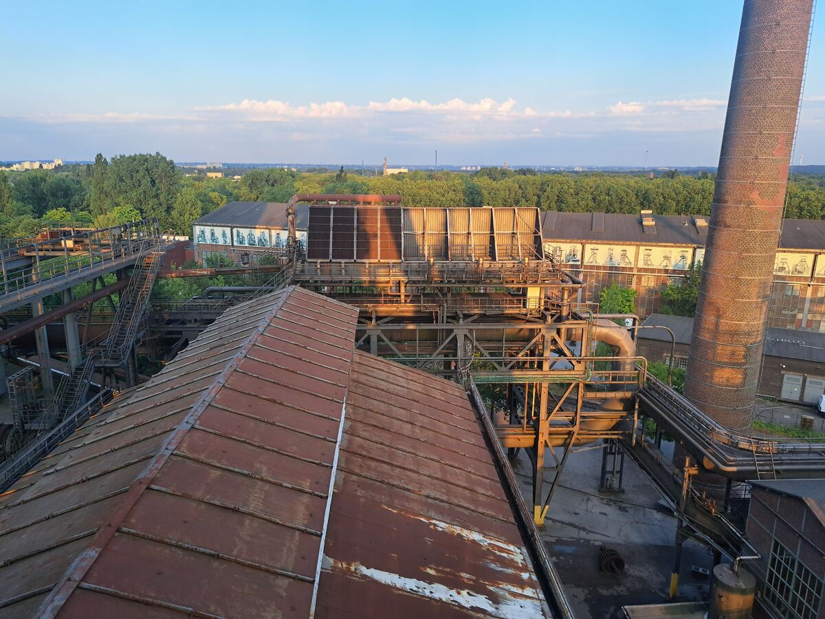 Een bovenaanzicht van een voormalig industrieel complex met roestige daken, metalen constructies, pijpen en loopbruggen. Een hoge bakstenen schoorsteen is rechts zichtbaar. Op de achtergrond staan gebouwen met kunstwerken op de gevels, omringd door veel groen. De lucht is blauw met witte wolken en de structuren worden beschenen door warm avondlicht rond 20:53 uur.