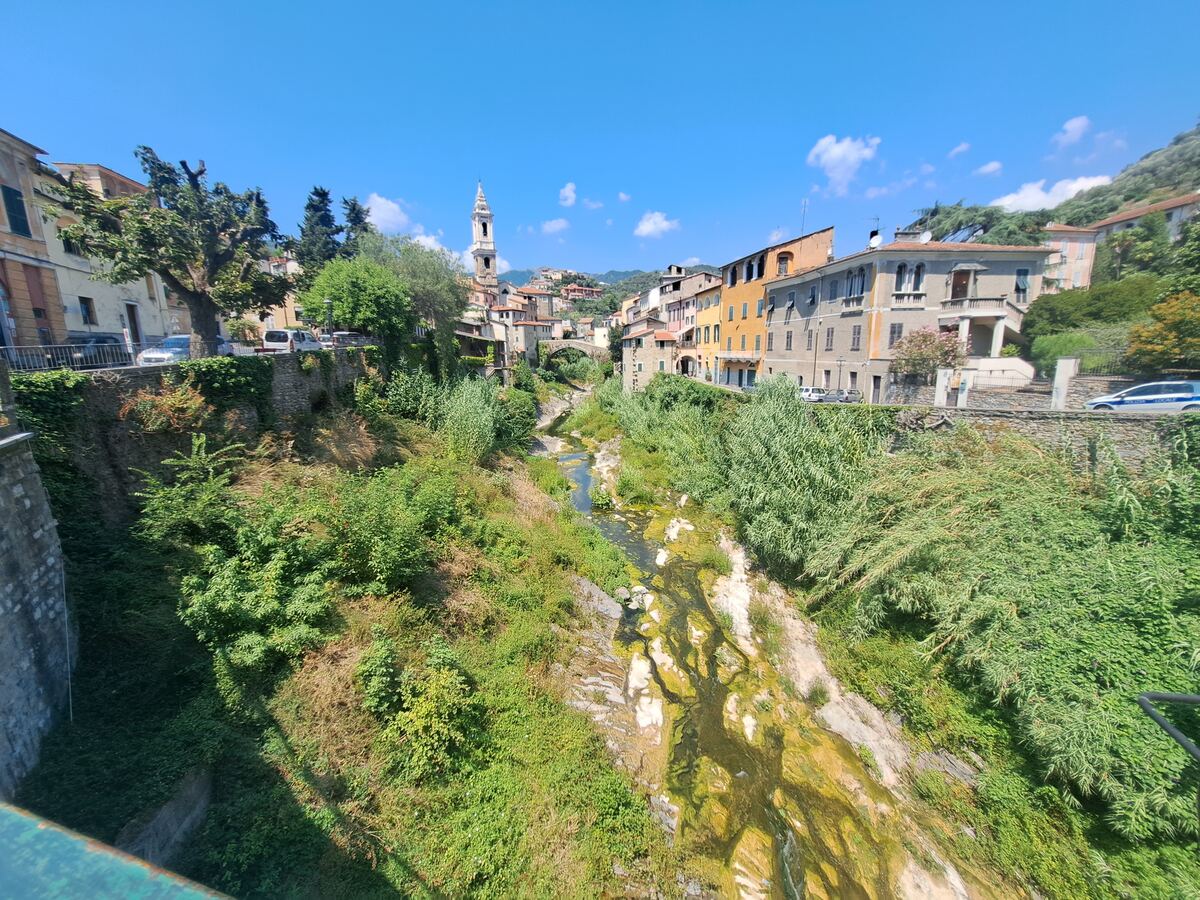 De foto toont een panoramisch uitzicht over een Italiaans dorp, gezien vanaf een brug. In het midden ligt een deels droge rivierbedding met rotsen en groenige algen, omzoomd door dichte groene begroeiing. Aan weerszijden van de rivier staan traditionele, kleurrijke gebouwen, variërend van okergeel tot lichtgrijs, langs wegen waar enkele auto's geparkeerd staan. Op de rechterweg is een witte auto met 'POLIZIA LOCALE' te zien. Een opvallende kerk met een hoge klokkentoren en spits domineert de sky