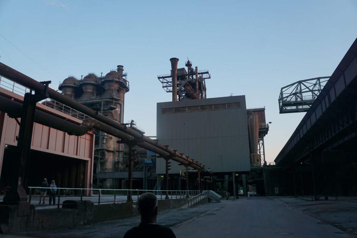 De foto toont een uitgestrekt industrieel landschap onder een helderblauwe avondlucht. Op de voorgrond is de rug van een persoon te zien die naar de industriële structuren kijkt. Diverse grote pijpleidingen lopen horizontaal door het midden van het beeld. Links staan imposante, roestbruine hoogovens met complexe constructies en ladders. Rechts daarvan staat een groot, grijs gebouw met een plat dak en daarbovenop meer industriële installaties. Rechtsboven is een deel van een metalen brugconstruct